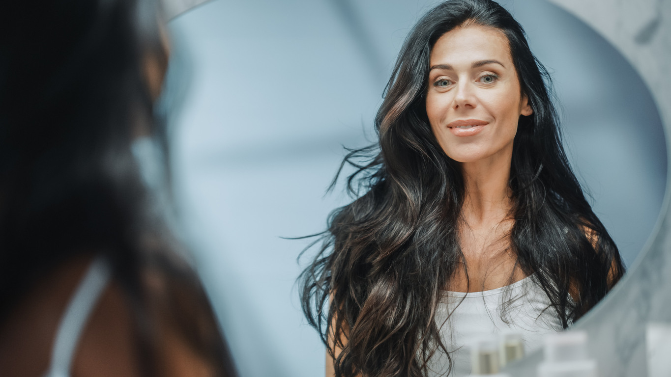 Beautiful Caucasian Middle Aged Woman Looks into Bathroom Mirror, Enjoys Her Looks. Concept for Happiness, Wellbeing, Natural Beauty and Juvederm Voluma® Restores Cheek Volume